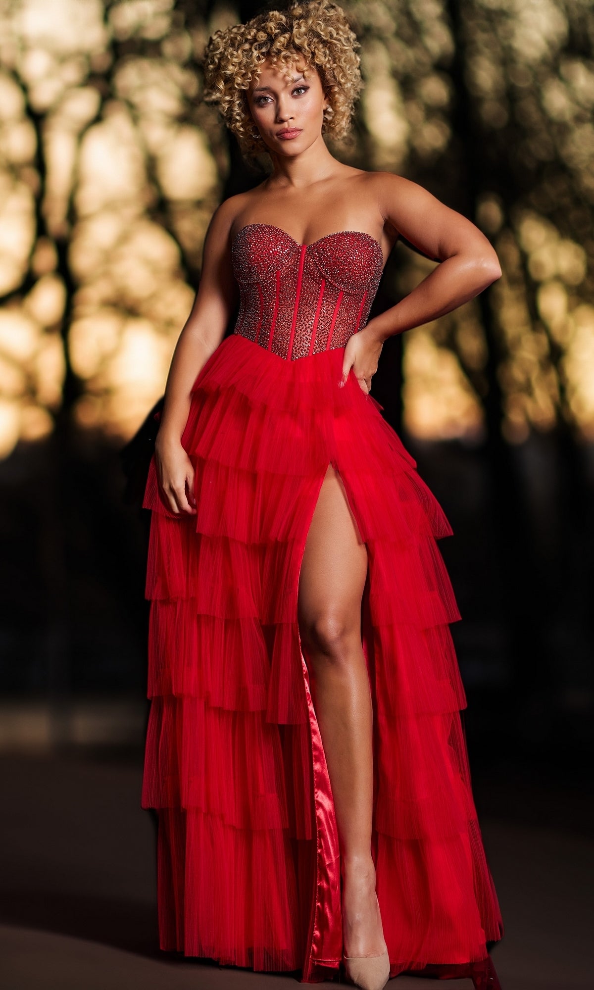 A woman stands outdoors in sunlight and trees wearing the Long Prom Dress 38090 by Jovani, featuring a strapless red tiered gown, embellished corset bodice, and high slit.